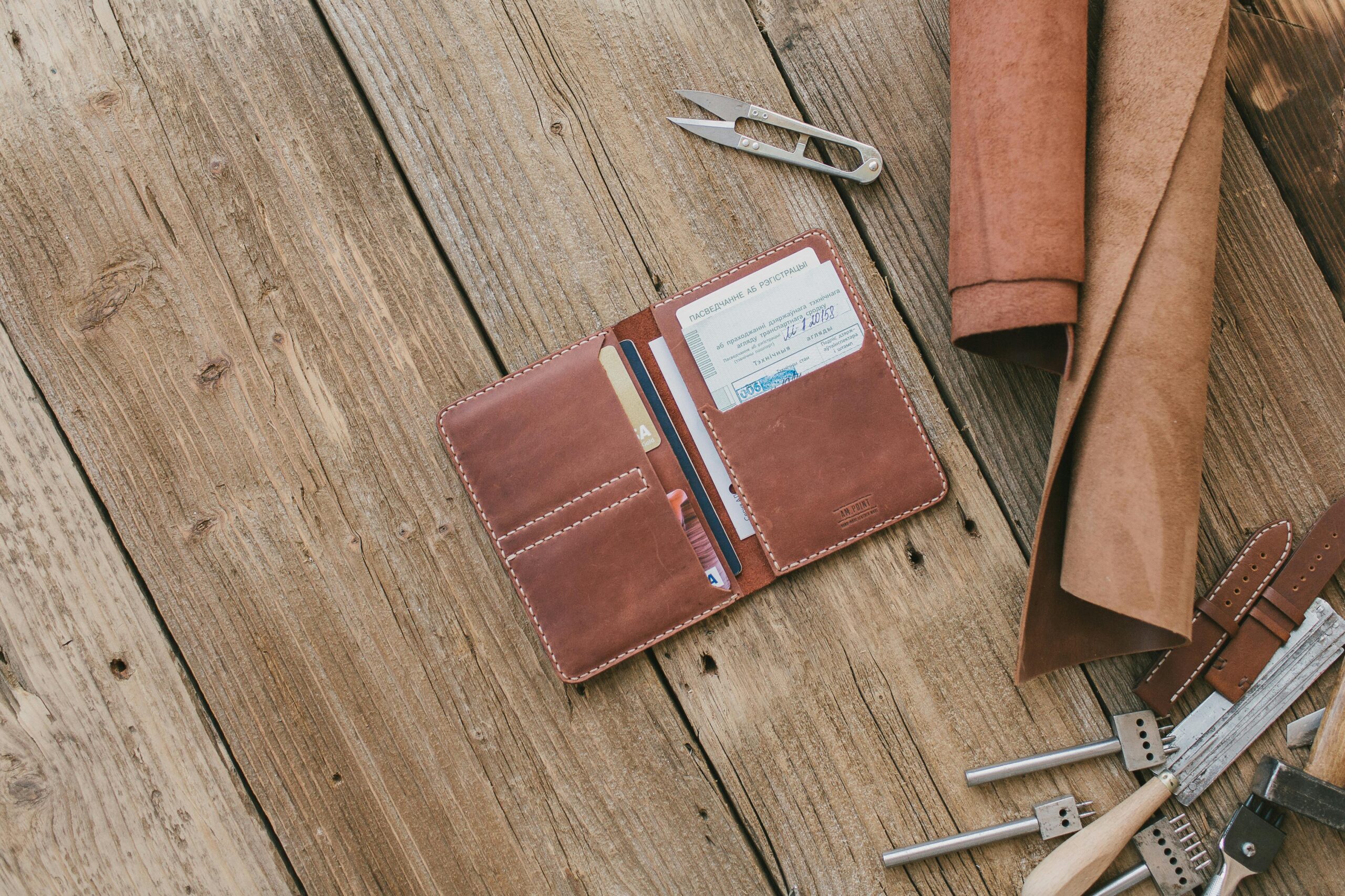 Brown leather wallet with accessories on wooden planks, featuring crafting tools and materials for leatherwork.